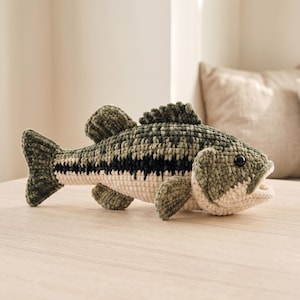 May include: A plush fish toy crafted with textured yarn in shades of olive green, dark forest green, and cream. The fish features a prominent black horizontal stripe along its side, a dorsal fin, pectoral fins, and a small, open mouth with a black eye.