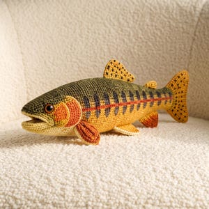 May include: A crocheted fish toy featuring a dark olive green back, a yellow underbelly, and a distinct red lateral stripe. The fish has a textured surface with dark vertical markings along its sides, orange gill covers, and a spotted tail fin.