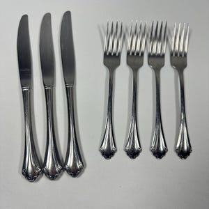 May include: A set of three silver-toned stainless steel dinner knives and four matching dinner forks. Each piece features a smooth, polished finish with a flared handle base that includes a subtle, embossed decorative scroll detail at the end.
