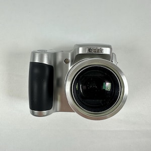 May include: A silver digital camera with a prominent black rubberized grip on the left side. The front features a large circular lens assembly and the Kodak brand name printed in black lettering on the top center of the camera body.