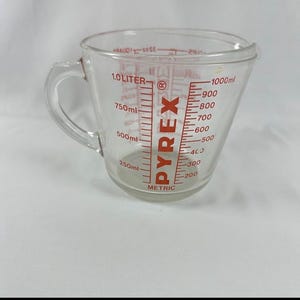 Pode incluir: Um copo medidor de vidro transparente com alça, apresentando marcações vermelhas para 1,0 litro e 1000 ml, além de incrementos métricos intermediários. A palavra PYREX está impressa verticalmente em vermelho com uma marca registrada.