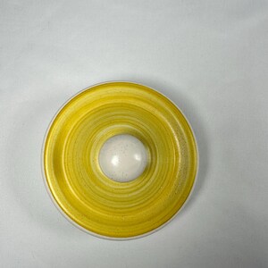 Pode incluir: Uma tampa de cerâmica redonda com um padrão de espiral amarelo brilhante e manchas subtis. O centro da tampa tem um botão esférico liso e elevado de cor branco sujo. O design é circular com uma borda bem definida.