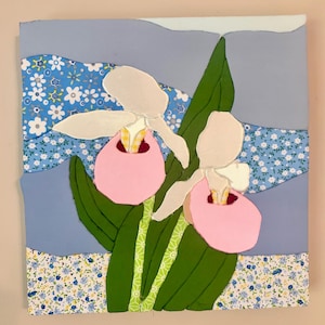 May include: A square fabric collage art piece featuring two pink and white lady slipper flowers with green stems and leaves. The background consists of layered blue and white floral patterned fabric strips and solid light blue fabric sections.