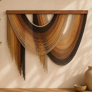 May include: A wall hanging featuring multiple strands of yarn in shades of black, brown, tan, and cream, suspended from a horizontal wooden bar. The yarn is arranged in overlapping, curved layers with a row of small, light-colored wooden beads.