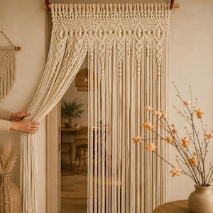 May include: A cream-colored macrame door curtain featuring an intricate knotted pattern at the top with diamond-shaped motifs and long, vertical hanging fringe strands suspended from a wooden rod, creating a textured, woven room divider.