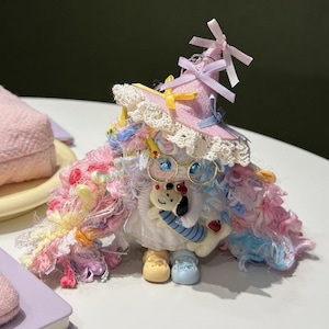 Op de afbeelding: Een pop met een lichtroze puntmuts met kanten rand en linten, voorzien van volumineus, veelkleurig garenhaar in tinten roze, blauw en wit. De pop draagt een goudkleurige bril en houdt een klein wit hondje vast als figuurtje.