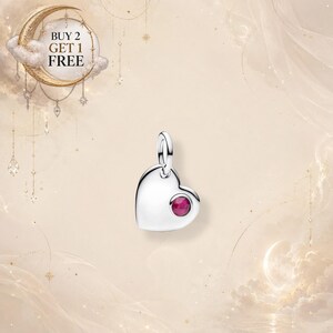 Puede incluir: Un dije colgante en forma de corazón de plata con una pequeña piedra preciosa redonda de color rosa intenso engastada en la parte inferior derecha. El dije incluye una pequeña argolla en la parte superior. El texto "BUY 2 GET 1 FREE" es visible.
