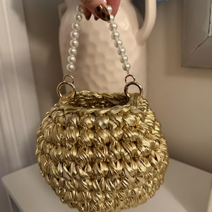 May include: A round, gold-colored crocheted handbag featuring a thick, textured metallic yarn construction. The bag includes a handle made of white, spherical faux pearls attached to the main body with gold-toned metal rings.