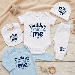 May include: A baby clothing set including a white short-sleeve bodysuit, a light blue long-sleeve bodysuit, white pants, a white bib, and a white beanie. Each piece features the dark blue text Daddy's mini me with heart and star graphics.