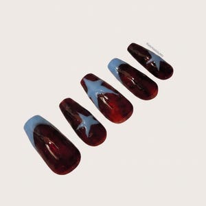 May include: A set of five press-on nails with a deep burgundy, semi-translucent base. Each nail features a light blue star design or a light blue French tip. The text RelicNailsbyMo is printed in small black letters on the smallest nail.