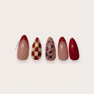 May include: Five almond-shaped press-on nails featuring varied designs: a red French tip, a red and white checkered pattern, a nude base with black X and O symbols, a red French tip, and a solid red nail. The text RelicNailsbyMo is visible.