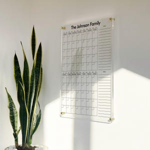 Pode incluir: Um calendário de parede em acrílico transparente com o texto The Johnson Family no topo. O calendário inclui duas grelhas mensais com os dias da semana e uma secção vertical de notas no lado direito, montado com quatro suportes dourados.