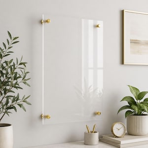 May include: A rectangular clear acrylic wall-mounted dry erase board featuring four polished gold-tone metal standoffs at each corner. The transparent panel is mounted against a neutral wall, providing a sleek surface for writing notes.
