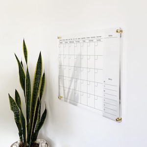 May include: A clear acrylic wall-mounted calendar featuring a grid for days of the week, a monthly header, a notes section, and a weekly menu list. The board is secured with four gold-toned metal standoffs at each corner, displaying black text.