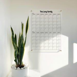 Könnte beinhalten: Ein Wandkalender aus klarem Acryl mit dem aufgedruckten Text The Long Family oben. Der Kalender verfügt über zwei Monatsraster mit Spalten von Montag bis Sonntag und ist mit vier goldfarbenen Wandabstandshaltern befestigt.