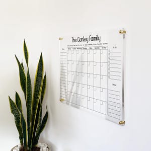 Könnte beinhalten: Ein Wandkalender aus klarem Acryl mit der Aufschrift The Conley Family, einem Monatsraster, einem Notizbereich, einer Aufgabenliste und einem Menübereich. Die Tafel ist mit vier goldenen Wandabstandshaltern an einer weißen Wand befestigt.