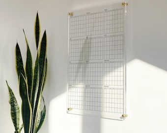 May include: A clear acrylic wall calendar featuring a twelve-month grid layout with black printed text for each month and day. The rectangular panel is mounted with four gold-toned metal standoffs at the corners, providing a floating effect.