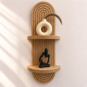 May include: A light wood wall shelf with an arched top and vertical grooved detailing. The shelf features two horizontal tiers. A cream-colored donut-shaped vase with a dried grass stem sits on the top shelf, and a black abstract figurine sits below.