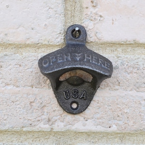 May include: A cast iron bottle opener mounted on a brick wall. The opener is dark gray with the words "OPEN HERE" and "USA" cast into the metal. A small hole is at the top for mounting.