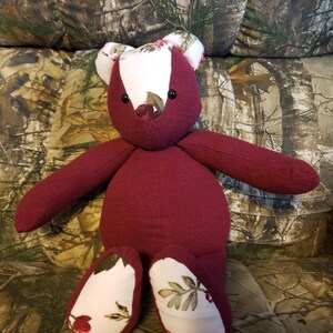 May include: A stuffed bear toy constructed from solid maroon ribbed fabric and white fabric featuring a floral pattern with red flowers and green leaves. The bear has two small black circular eyes and a stitched nose, with floral fabric on the ears and feet.