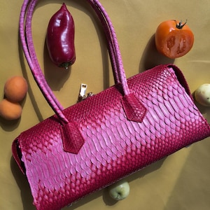 May include: A cylindrical handbag featuring a vibrant pink and magenta ombre scale pattern. The bag has two matching pink tubular handles and a gold-toned zipper closure. The exterior texture mimics a snakeskin finish with overlapping scales.