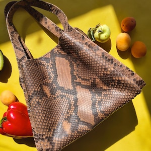 Peut inclure: Un sac fourre-tout avec un motif peau de serpent marron et noir et deux anses. Le sac est posé sur une surface jaune vif à côté d'un poivron rouge, de trois petits fruits à noyau orange et d'une pomme verte avec une petite feuille.