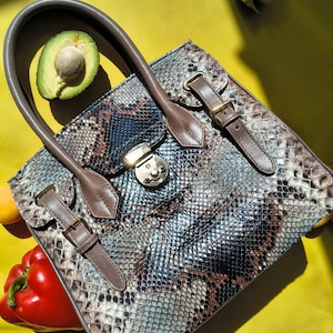 Op de afbeelding: Een gestructureerde handtas met een slangenprint aan de buitenkant in grijze, bruine en zwarte tinten. De tas is voorzien van een bruin handvat, een centrale goudkleurige metalen sluiting en twee decoratieve bruine riemen met gespen.