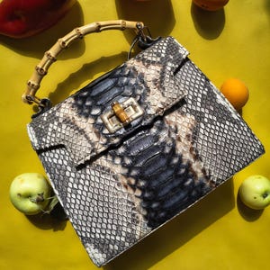 May include: A structured handbag featuring a snakeskin pattern in shades of gray, black, and white. The bag includes a curved bamboo top handle and a rectangular bamboo turn-lock closure on the front flap, showcasing a detailed scale texture.