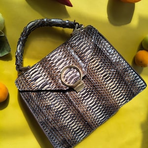 May include: A structured handbag featuring a textured, snakeskin-patterned exterior in shades of brown, gray, and cream. The bag includes a curved top handle, a triangular flap closure, and a prominent circular gold-toned metal clasp on the front.