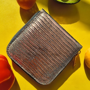 Puede incluir: Una cartera de forma cuadrada con un acabado en bronce metálico y un patrón texturizado de piel de lagarto. La cartera está asegurada con una cremallera en el borde superior e incluye un pequeño tirador de metal dorado en el lateral.