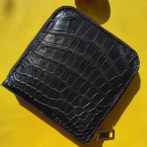 May include: A square black wallet featuring a textured crocodile skin pattern. The wallet has rounded corners and a metal zipper closure with a rectangular pull tab. The surface of the wallet displays a glossy finish with deep embossed scales.