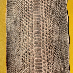 May include: A rectangular strip of metallic bronze snakeskin leather featuring a distinct central row of large, elongated scales flanked by smaller, tightly packed scales on both sides, showcasing a natural reptilian texture and sheen.