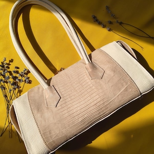 Könnte beinhalten: Eine rechteckige Handtasche mit einem Mittelteil in hellbeiger Eidechsenhaut-Optik, flankiert von glatten, cremeweißen Lederseiten. Sie verfügt über zwei passende, cremeweiße Schulterriemen aus Leder, die an der Oberseite befestigt sind.