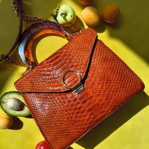 May include: A structured handbag with a textured, rust-colored snakeskin-patterned exterior, featuring a rounded tortoiseshell-style handle, a gold-toned chain strap, and a circular gold-toned metal clasp on the front flap closure.