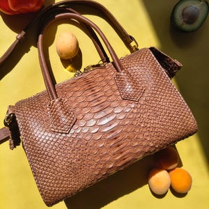 May include: A brown handbag with a textured snakeskin pattern, featuring two top handles and a detachable shoulder strap. The bag has a rectangular shape with gold-toned hardware accents, including a zip closure along the top edge.