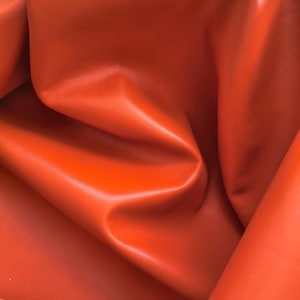 May include: A vibrant orange leather hide with a smooth, matte finish. The material is folded and draped to create deep, soft curves and shadows, highlighting the consistent, saturated colour and the supple texture of the surface.