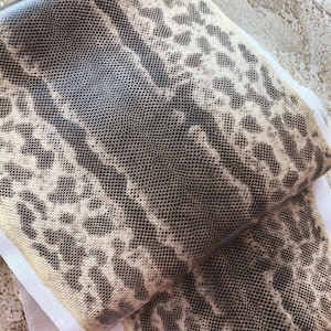 May include: A folded piece of fabric featuring a dark brown and cream snakeskin pattern with a textured, scale-like appearance. The material has a distinct reptilian print with alternating dark and light sections, showing a white underside.