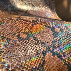 Könnte beinhalten: Ein Stück texturiertes Material mit einem sich wiederholenden Schlangenhautmuster in Braun-, Schwarz- und Beigetönen. Die Oberfläche weist ein ausgeprägtes irisierendes Finish auf, das lebendige Regenbogenfarben wie Grün, Blau und Gold reflektiert.
