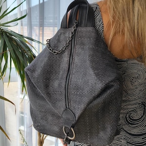 Puede incluir: Una mochila gris oscuro con un patrón de chevrón texturizado, un cierre de cremallera vertical central y un asa superior de aspecto de cuero negro. La bolsa incluye una cadena decorativa de metal oscuro y una anilla metálica en la base.