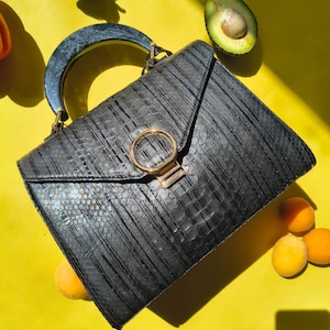 May include: A black handbag with a textured snakeskin pattern, a semi-circular dark handle, and a gold-toned circular clasp on the front flap. The bag features a structured design with vertical lines and a metallic closure mechanism.
