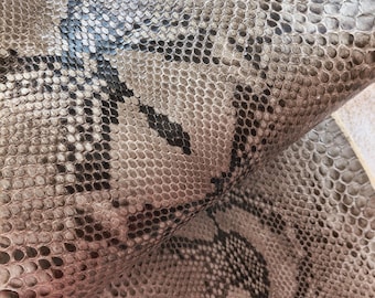 May include: A piece of flexible, textured snakeskin leather featuring a natural scale pattern in shades of gray, beige, and black. The material is folded over to reveal the detailed, overlapping scale texture and the organic, dark diamond-shaped markings.