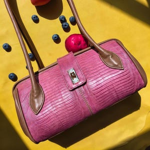 Könnte beinhalten: Eine pinke Handtasche mit texturiertem Eidechsenhaut-Muster, braunen Lederbesätzen und zwei braunen Schulterriemen. Die Tasche verfügt über einen zentralen Klappenverschluss mit einem goldenen Drehverschluss vor einem hellgelben Hintergrund.