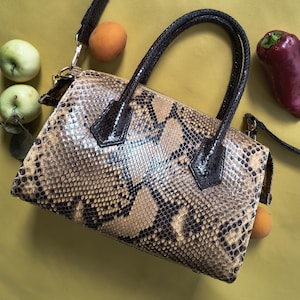 May include: A structured handbag featuring a snakeskin pattern in shades of tan, beige, and dark brown. The bag includes two dark brown top handles, a detachable shoulder strap with gold-tone hardware, and a top zipper closure.