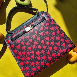 May include: A black handbag featuring a repeating pattern of small red hearts and dots. The bag has a top handle, a thin shoulder strap, and a horizontal black strap across the front secured with a metallic gold-toned rectangular clasp closure.