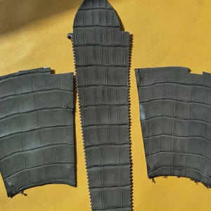 May include: Three pieces of dark gray material with a textured crocodile skin pattern. The set includes one long, narrow, pointed central piece and two wider, trapezoidal side pieces, all featuring a repeating rectangular scale design.