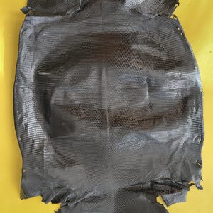 Può includere: Una pelle di lucertola grigio antracite scuro caratterizzata da un motivo a scaglie fine e uniforme su tutta la superficie. Il cuoio è conciato e appiattito, mostrando una trama simmetrica con piccole scaglie rettangolari di varie dimensioni.
