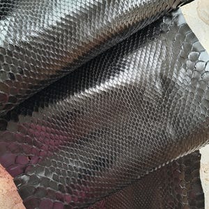May include: A piece of black snakeskin leather featuring a glossy, reflective finish and a distinct, repeating scale pattern. The material is folded over itself, showcasing the texture and the uniform arrangement of the individual scales.