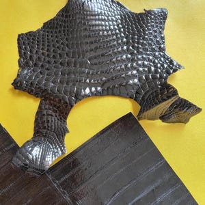 May include: A dark brown, glossy, textured piece of leather with a distinct scale pattern resembling crocodile skin, positioned above a separate, rectangular piece of dark brown leather featuring a linear, ribbed texture.
