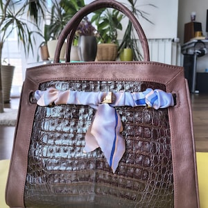 May include: A brown handbag featuring a dark brown crocodile-embossed center panel and smooth brown leather trim. A light purple, blue, and white silk scarf is tied around the front, secured by a central gold-toned metal clasp and two handles.