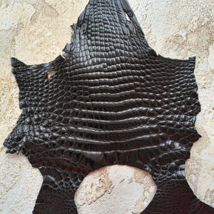 May include: A dark black, irregularly shaped piece of leather featuring a prominent, raised, and glossy crocodile-style scale texture across its entire surface, with visible natural edges and small perforations along the top section.